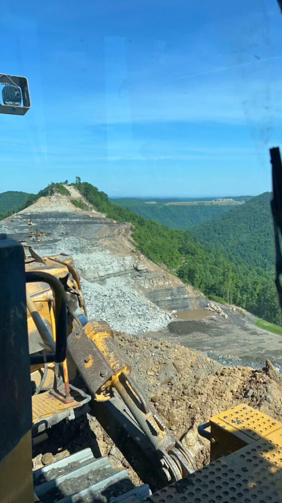 Excavator on a mountainside with a panoramic view of a cleared land area and lush green hills.